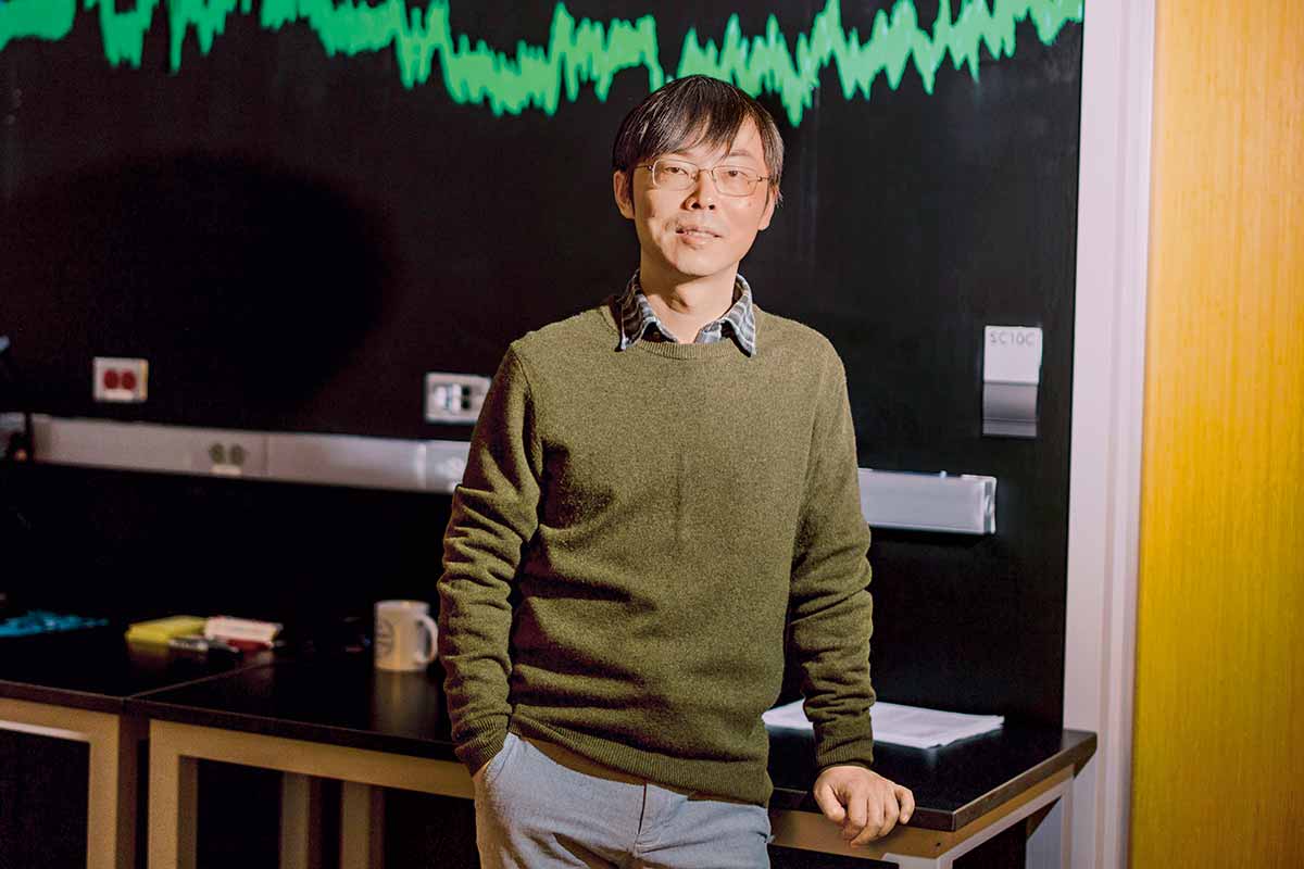 Liu leans against a desk with a bright green diagram behind them